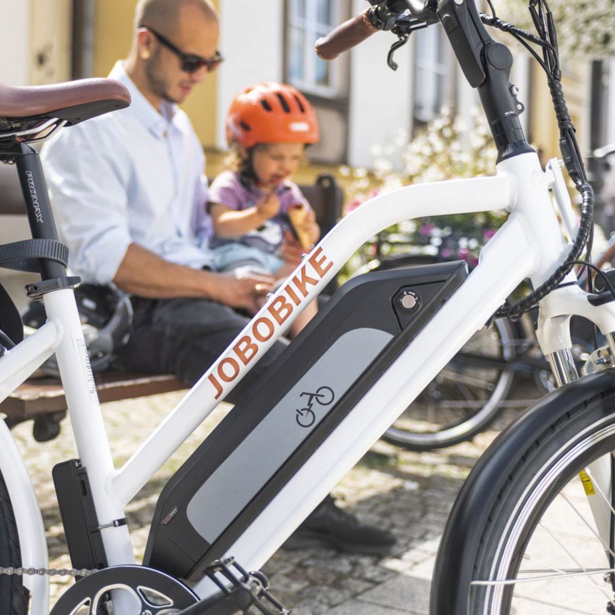 Vélo électrique blanc avec batterie, logo JOBOBIKE. Homme et enfant assis sur un banc en arrière-plan.