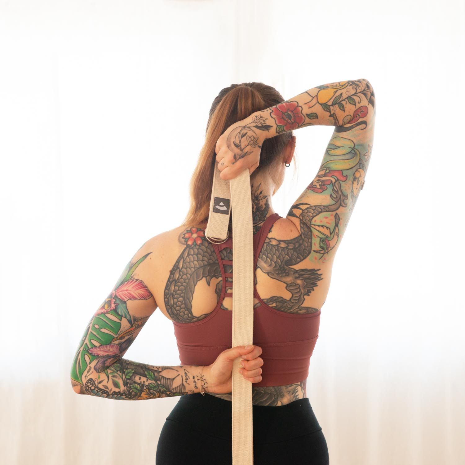 Femme tatouée tenant une sangle de yoga beige derrière le dos. Anneaux métalliques visibles.