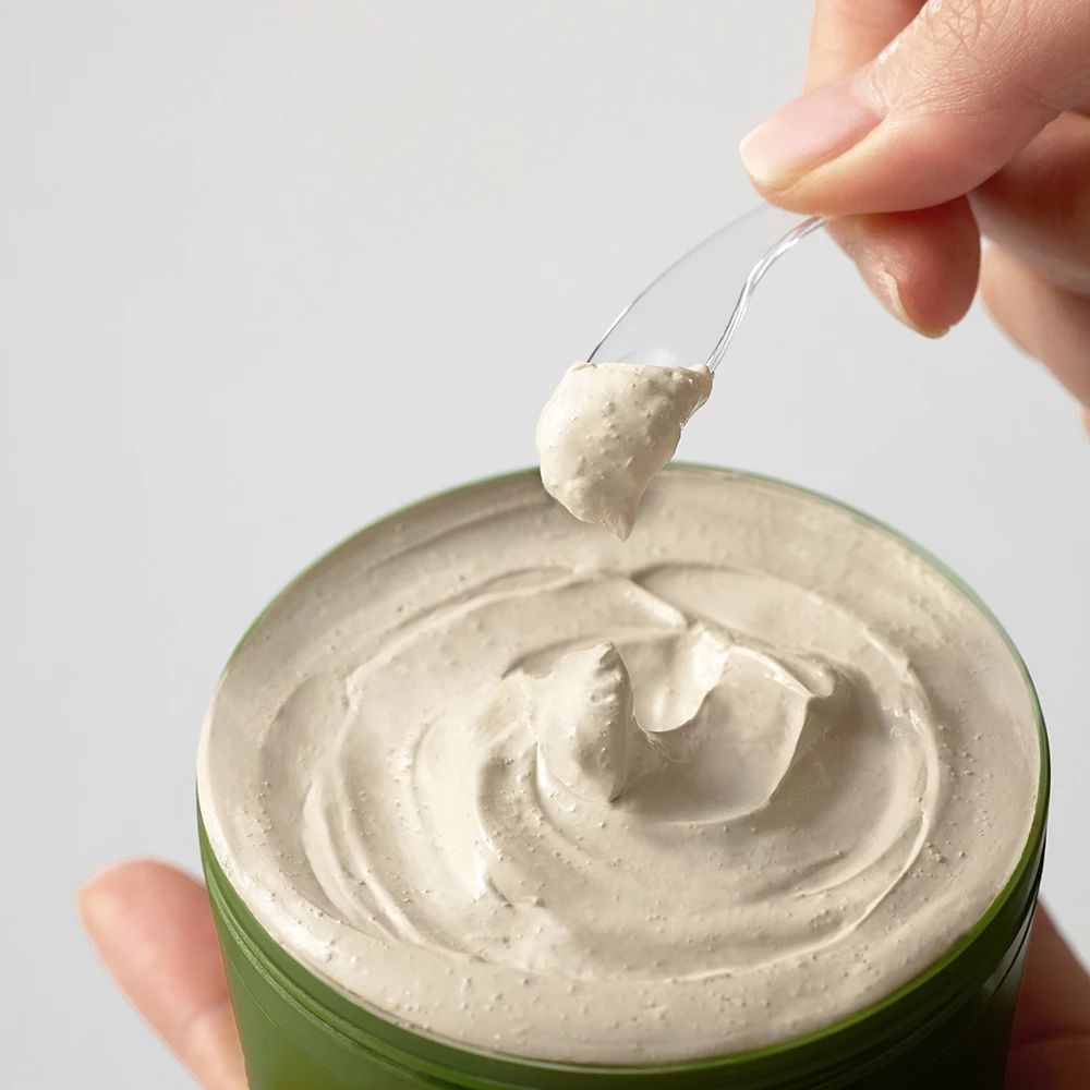 Groene pot met masker. Hand houdt lepel met masker. Crèmige, beige textuur. Pot open.