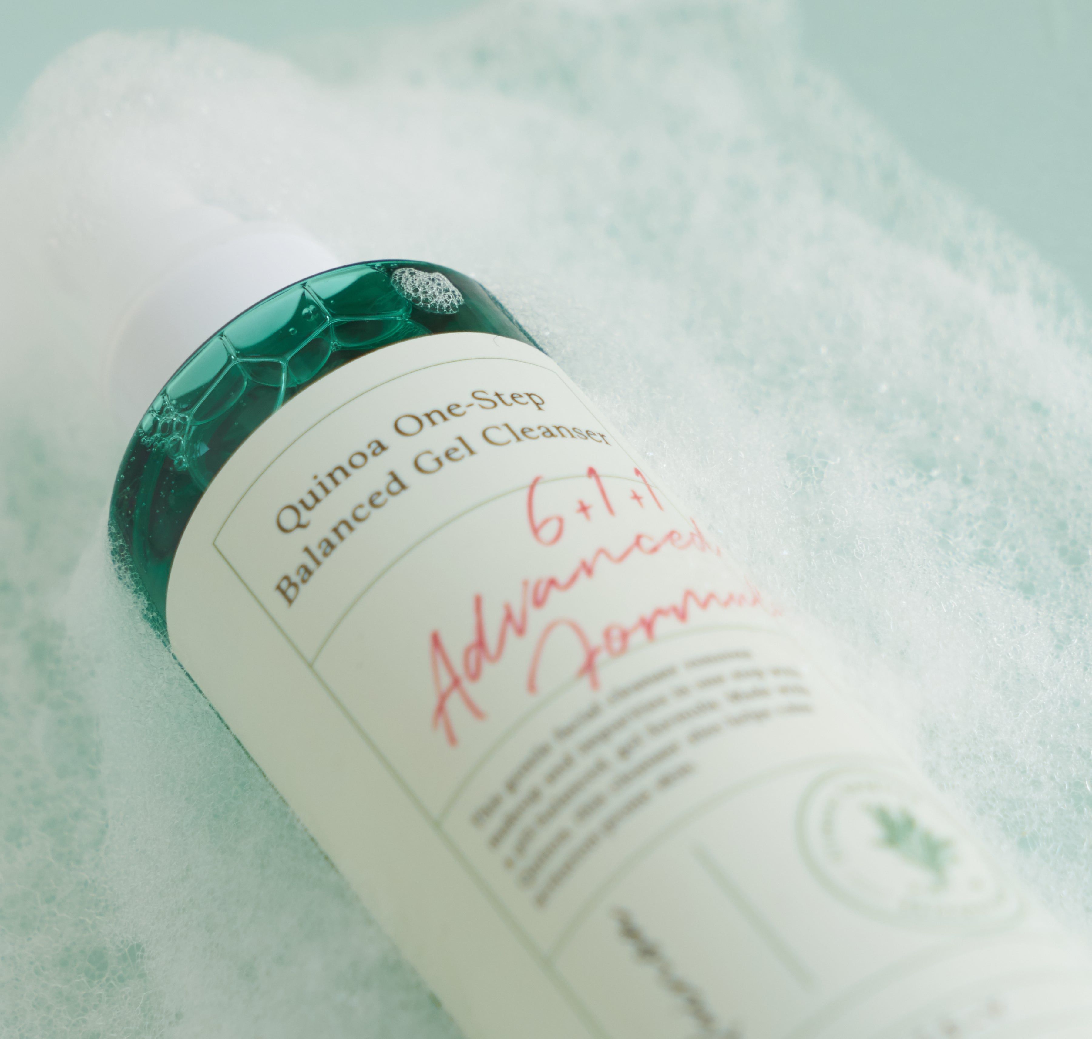 Close-up van een fles met schuim. Opschrift: AXIS-Y, Quinoa One-Step Balanced Gel Cleanser. Groen-wit etiket.