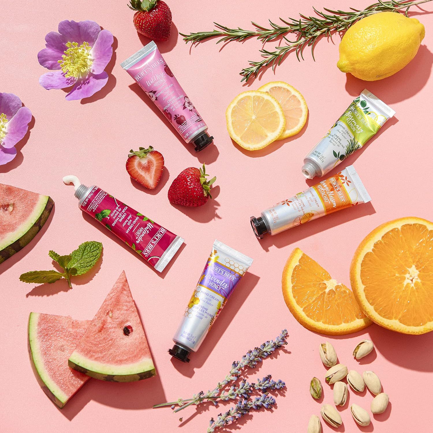 Meerdere handcrème tubes in verschillende kleuren en met fruitmotieven, omringd door fruit en bloemen.