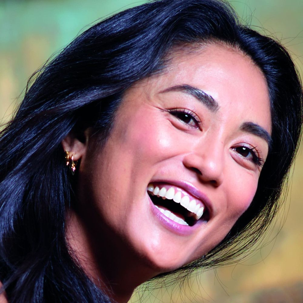 Portrait d'une femme souriante aux cheveux noirs. Elle porte des boucles d'oreilles et a le teint rosé. Gros plan.