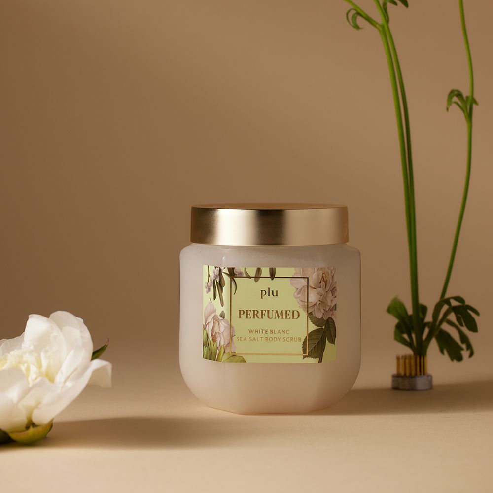 Pot en verre avec couvercle doré, à côté d'une fleur blanche. Étiquette avec nom du produit : PERFUMED White Blanc Sea Salt Body Scrub.