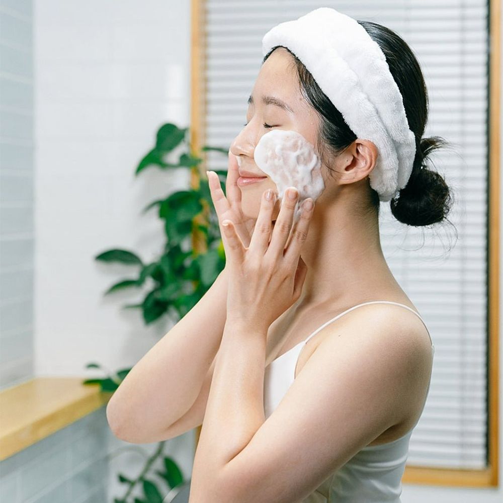 Femme avec bandeau blanc, appliquant de la mousse sur le visage. Mains sur le visage. Arrière-plan: salle de bain.