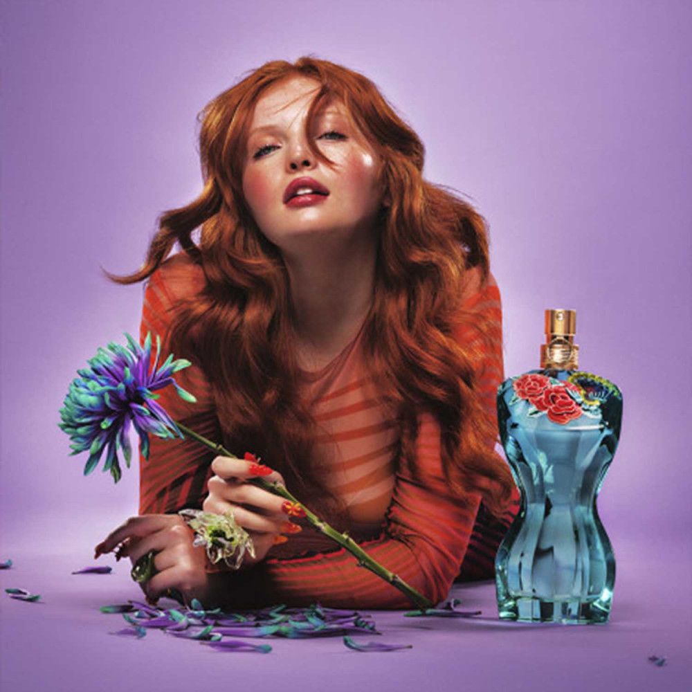 Femme aux cheveux roux et haut rouge. À côté, un flacon de parfum bleu en forme de corps féminin, décoré de fleurs.
