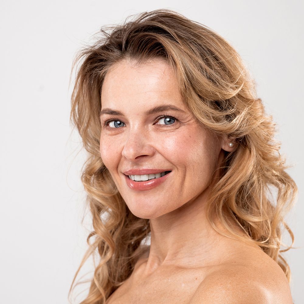 Portret van een lachende vrouw met blonde krullen. Ze kijkt recht in de camera. Lichte huidskleur.