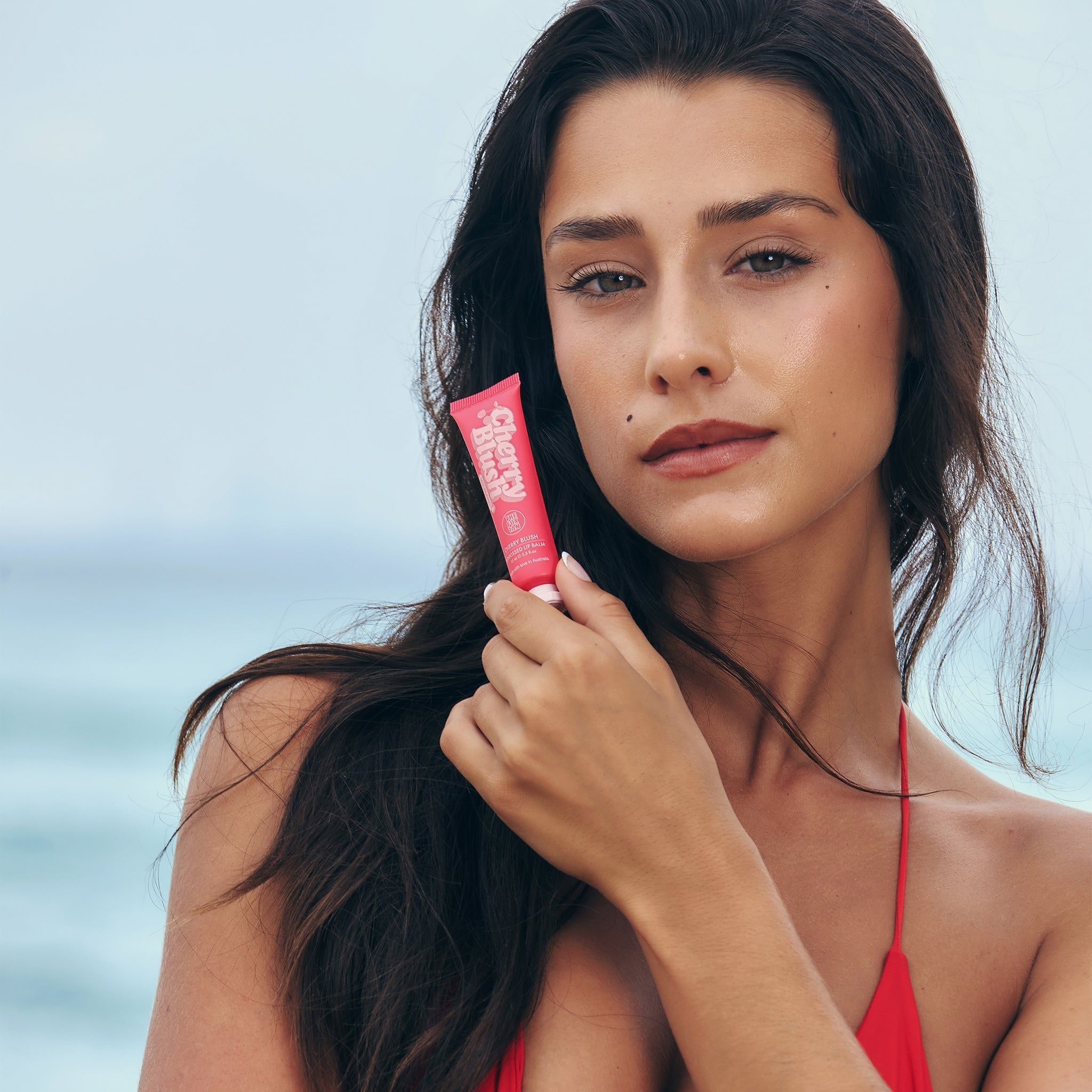 Femme tenant un tube 'Cherry Blush'. Nom du produit et marque visibles. Haut rouge. Arrière-plan plage.