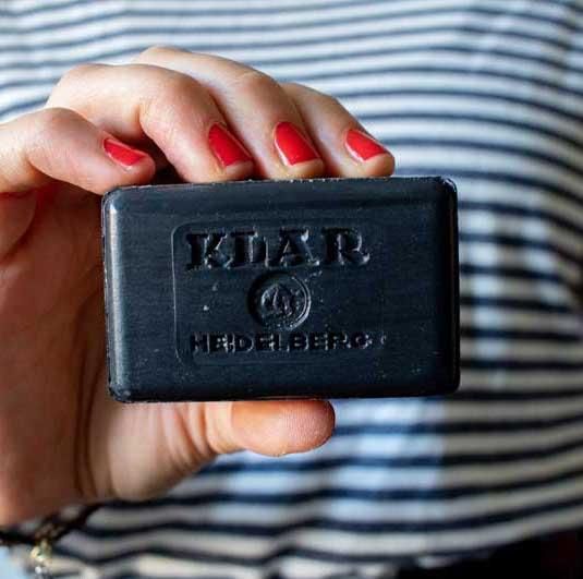 Hand houdt zwarte zeep vast. Zeep met reliëf Klar-logo en tekst. Rechthoekige vorm. Rode nagels.