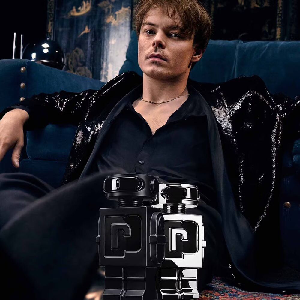 Homme assis sur un canapé, tenant deux flacons robots. Un argenté, un noir. Vêtements noirs.