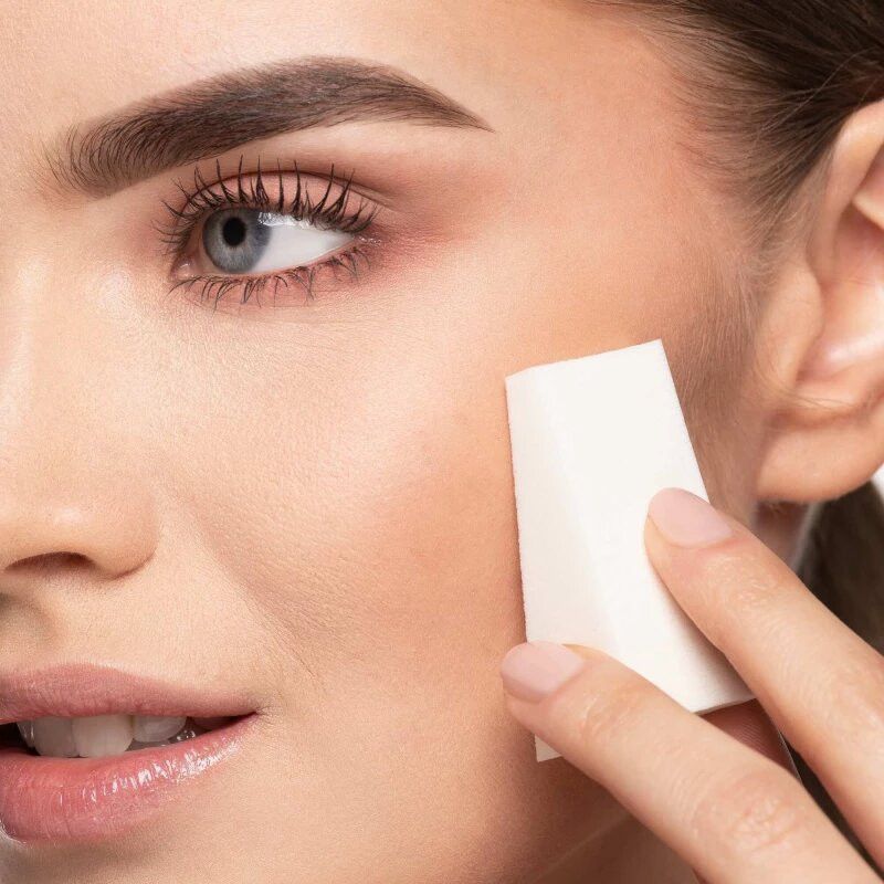 Une femme applique du fond de teint sur sa joue avec une éponge de maquillage blanche.