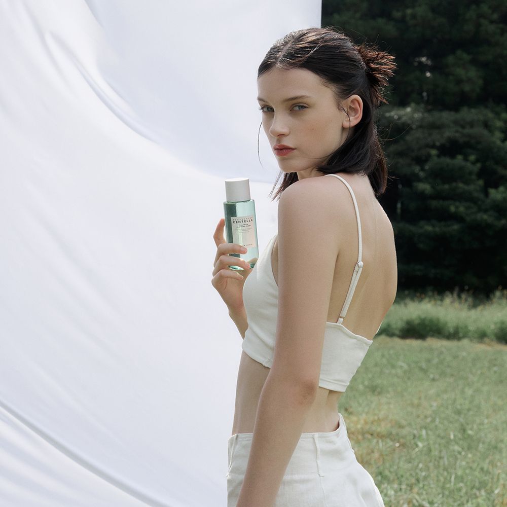 Vrouw houdt een groene fles vast. Fles met witte dop. Opschrift: MADAGASCAR CENTELLA. Merk: SKIN1004.