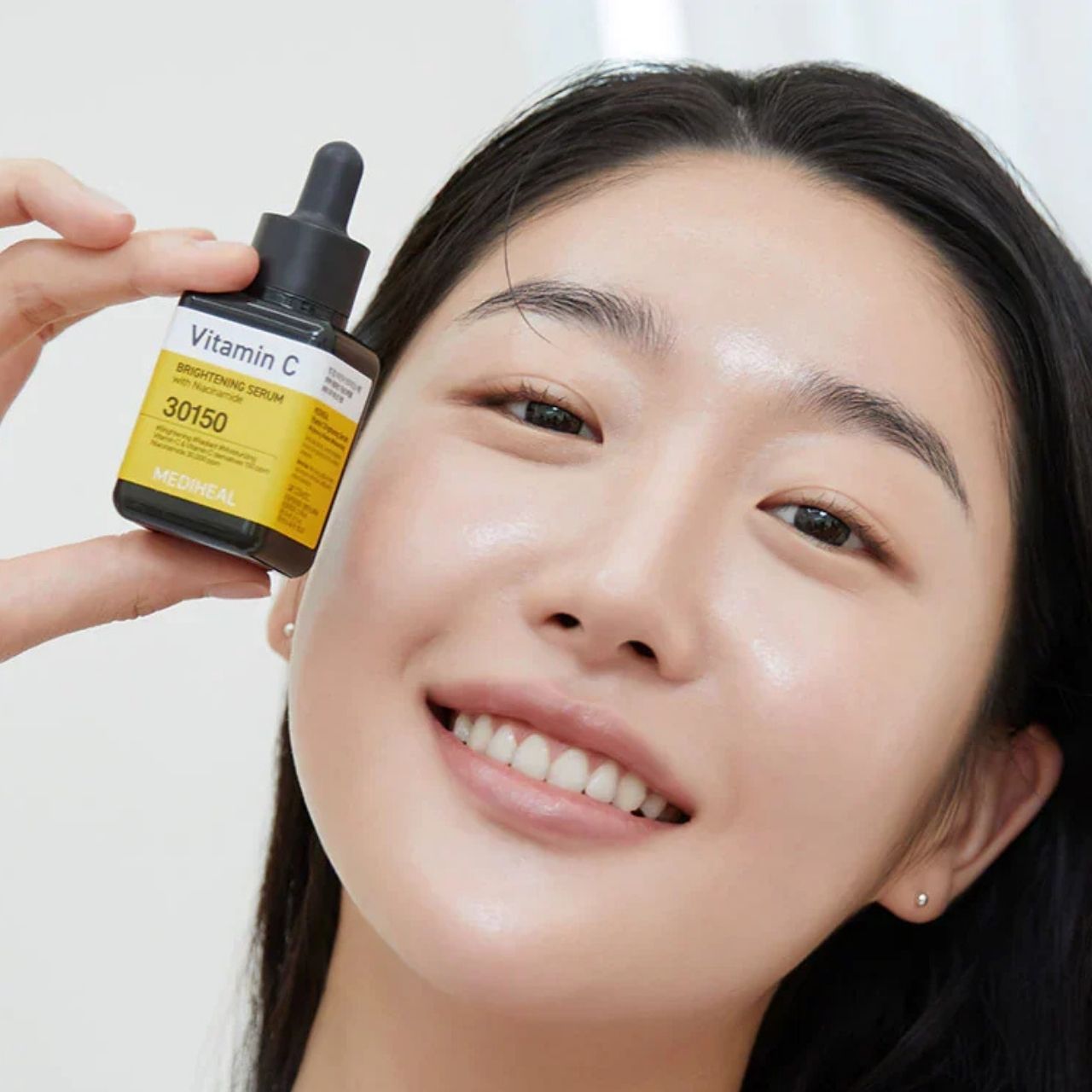 Une femme tient un flacon de sérum Mediheal Vitamine C devant son visage. Étiquette jaune avec le nom du produit.
