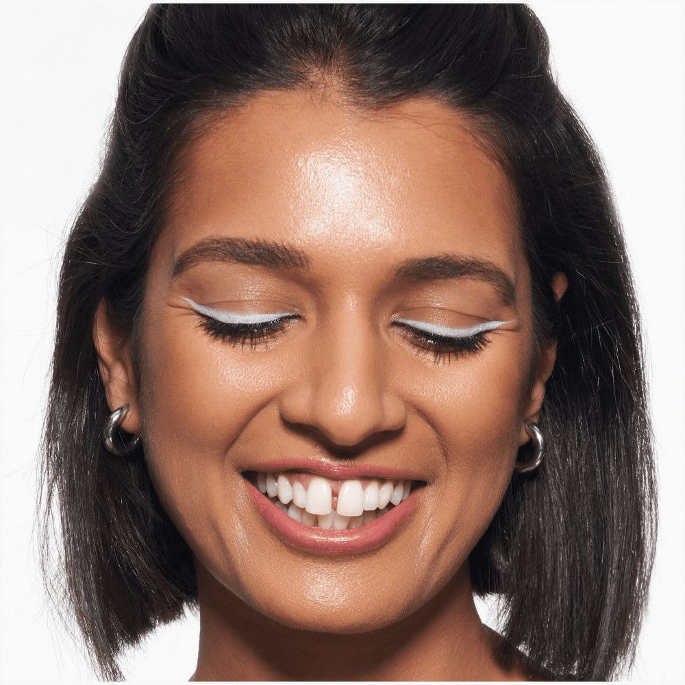 Close-up van een lachend gezicht met witte eyeliner. De vrouw heeft donker haar en draagt zilveren oorbellen.