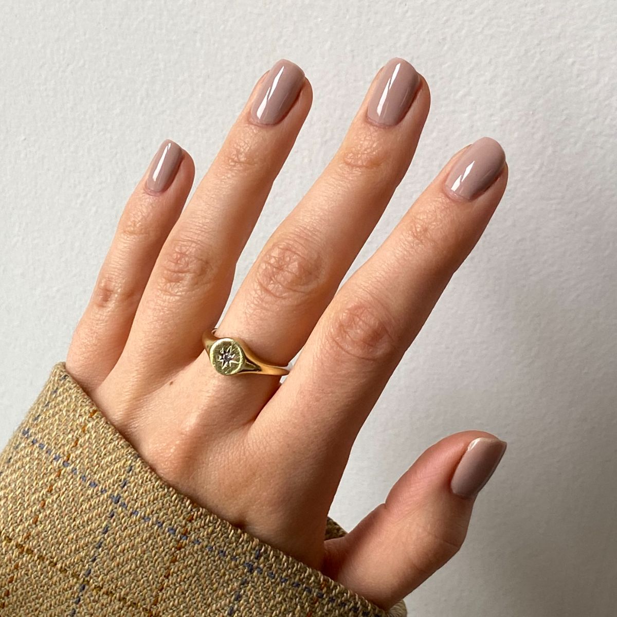 Main avec ongles peints en beige-brun. La personne porte une bague en or. La personne porte une veste.