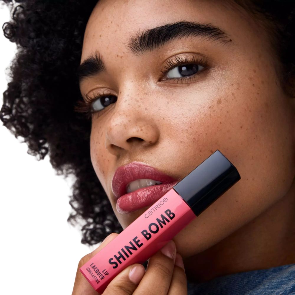 Femme avec rouge à lèvres rose. Emballage de rouge à lèvres en main. Inscription: SHINE BOMB. Marque: Catrice.