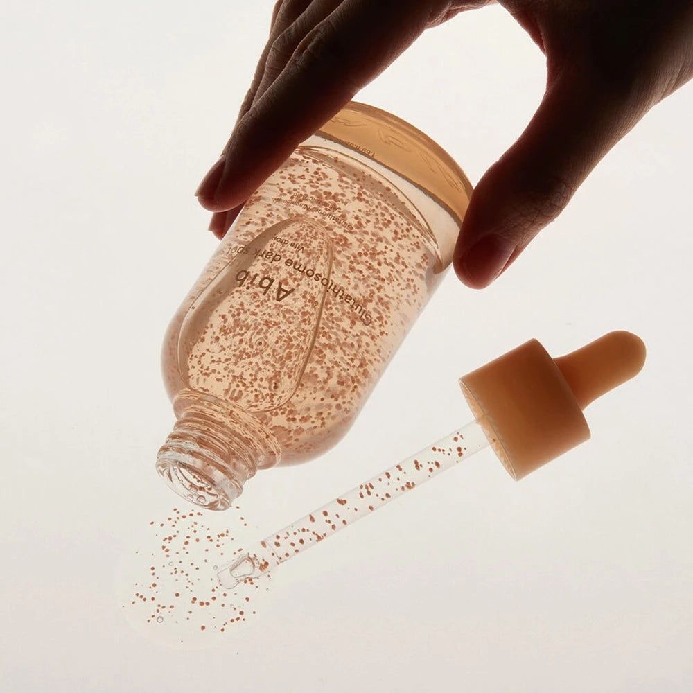 Flacon en verre avec sérum, billes roses, pipette. Une main tient le flacon. Le sérum est versé sur une surface.