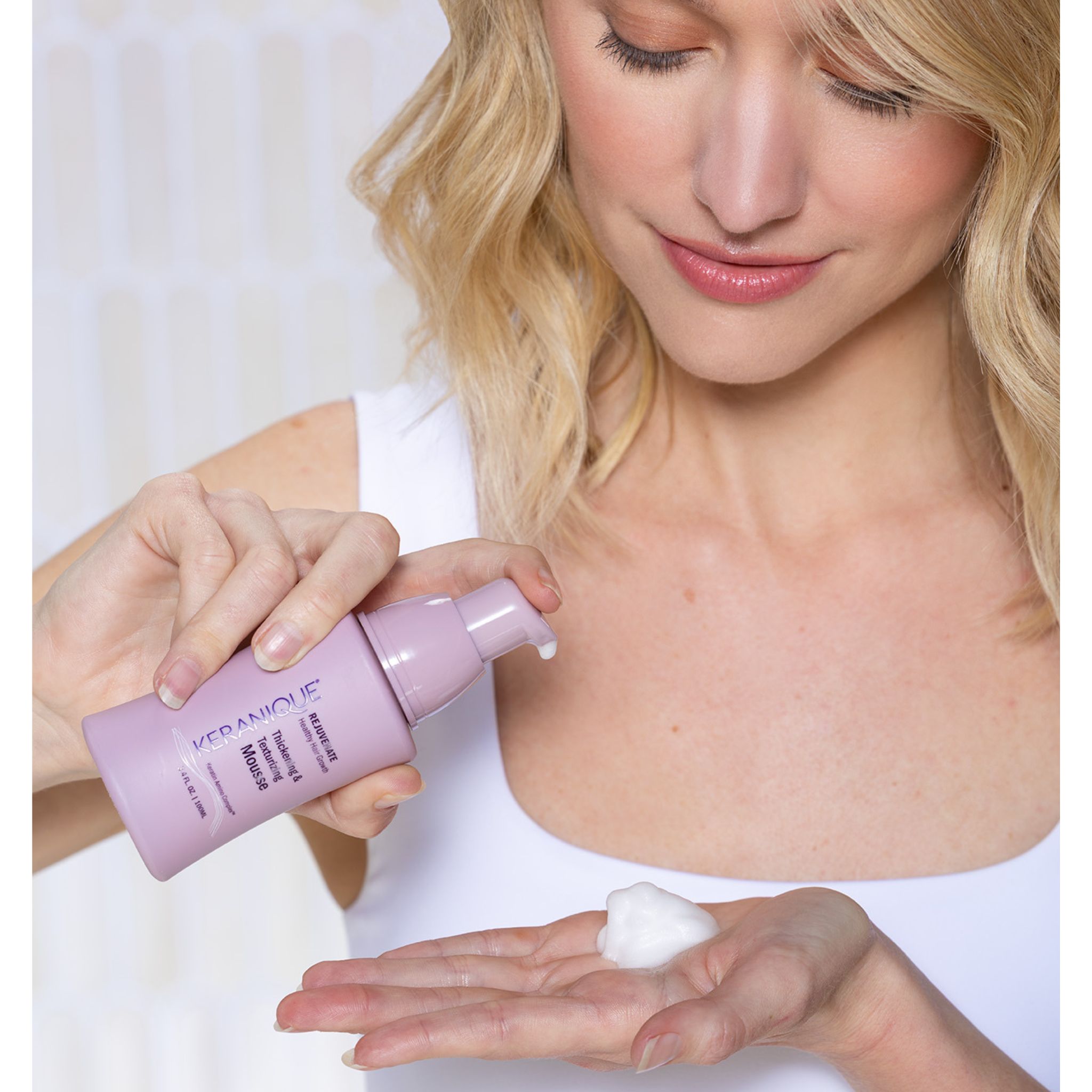 Vrouw houdt fles Keranique Thickening Mousse vast. Ze doseert mousse in haar handpalm. Lichtroze fles.
