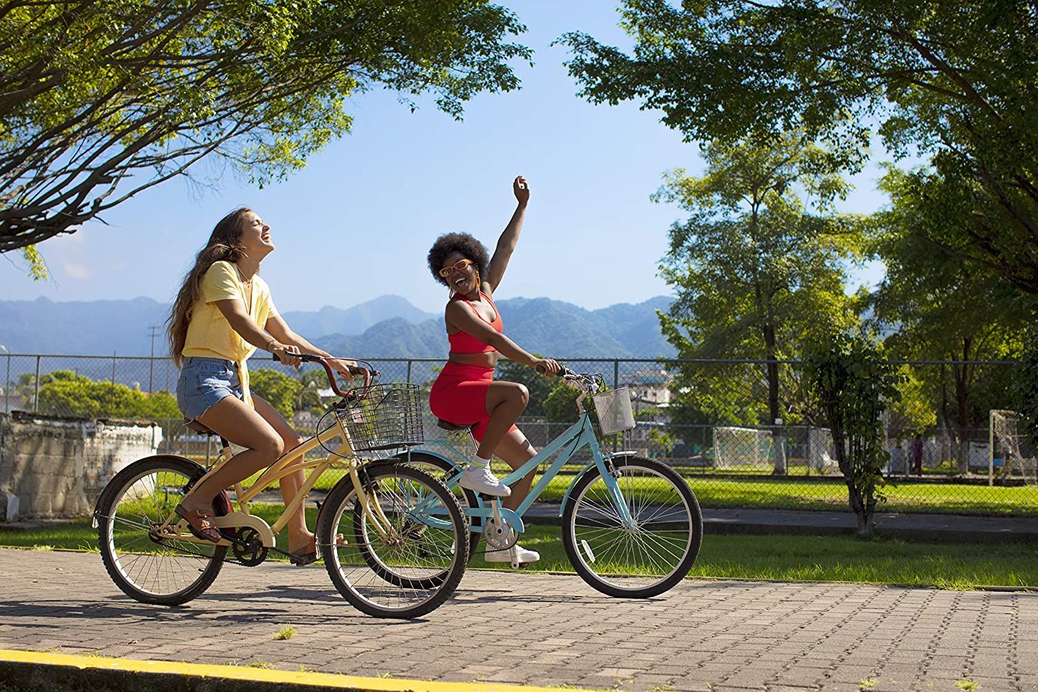 Deux jeunes femmes font du vélo. L'une porte une tenue rouge, l'autre un haut jaune. Soleil et arbres.