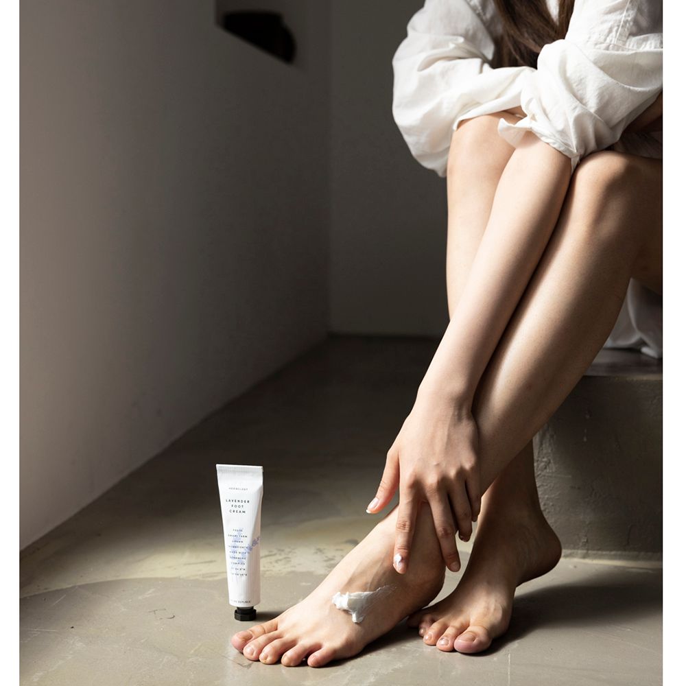 Persoon met voeten en voetcrèmetube. Crème wordt op voet aangebracht. Merk: NATURE REPUBLIC. Tekst: Herbology, Lavender Foot Cream.