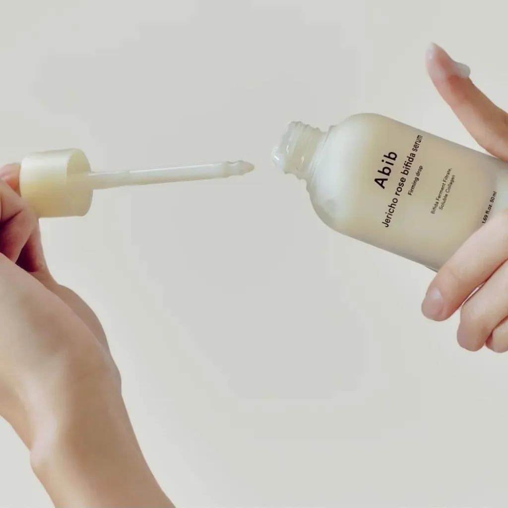 Handen houden een serumfles en pipet. Fles met opschrift: Abib Jericho Rose Bifida Serum Firming Drop. Pipet neemt serum op.