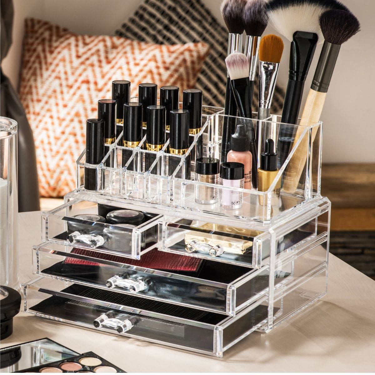 Transparante acryl make-up organizer met meerdere vakken en lade, gevuld met make-up en kwasten.