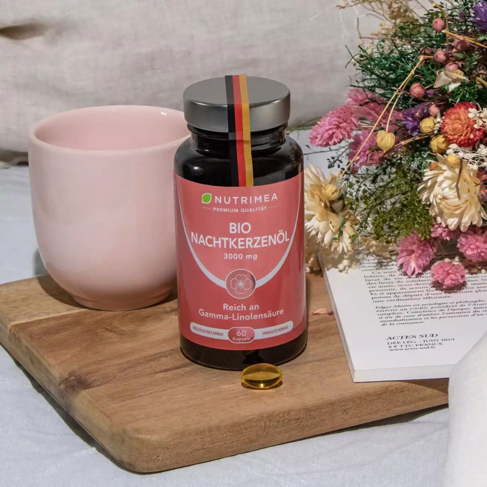 Fles met capsules op houten plank. Daarnaast een capsule en een kopje. Bloemenarrangement.