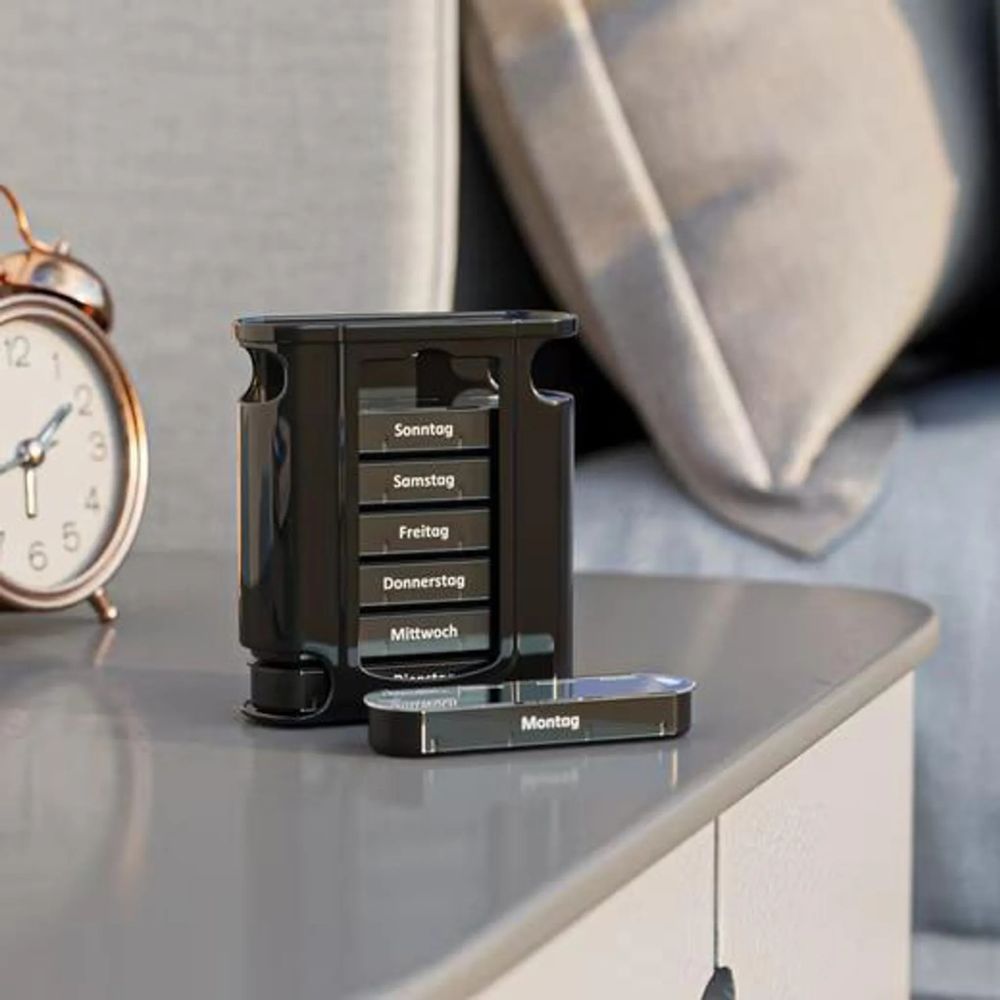 Zwarte pillendoos op een grijs oppervlak. Een verwijderd vak met de tekst Maandag. Op de achtergrond een klok en een kussen.