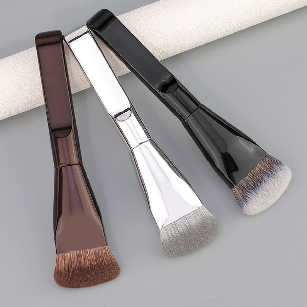 Trois pinceaux de maquillage plats avec des manches brillants marron, argent et noir.