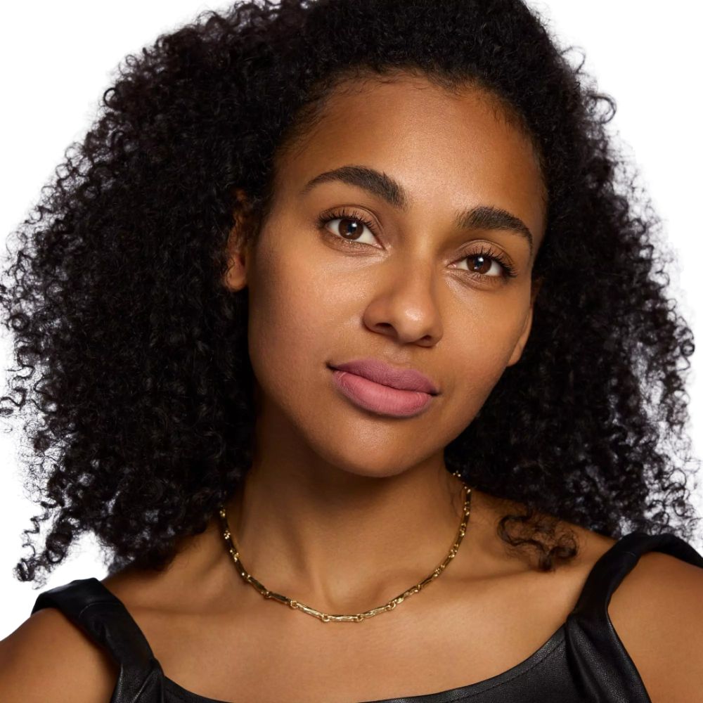 Portrait d'une femme portant du rouge à lèvres rose. Elle porte un collier doré et un haut noir. Ses cheveux sont bouclés.