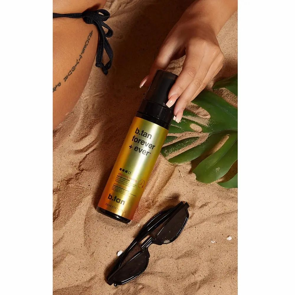 Fles zelfbruinende mousse op zand. Naast een hand, zonnebril en blad. Opschrift: b.tan forever + ever.