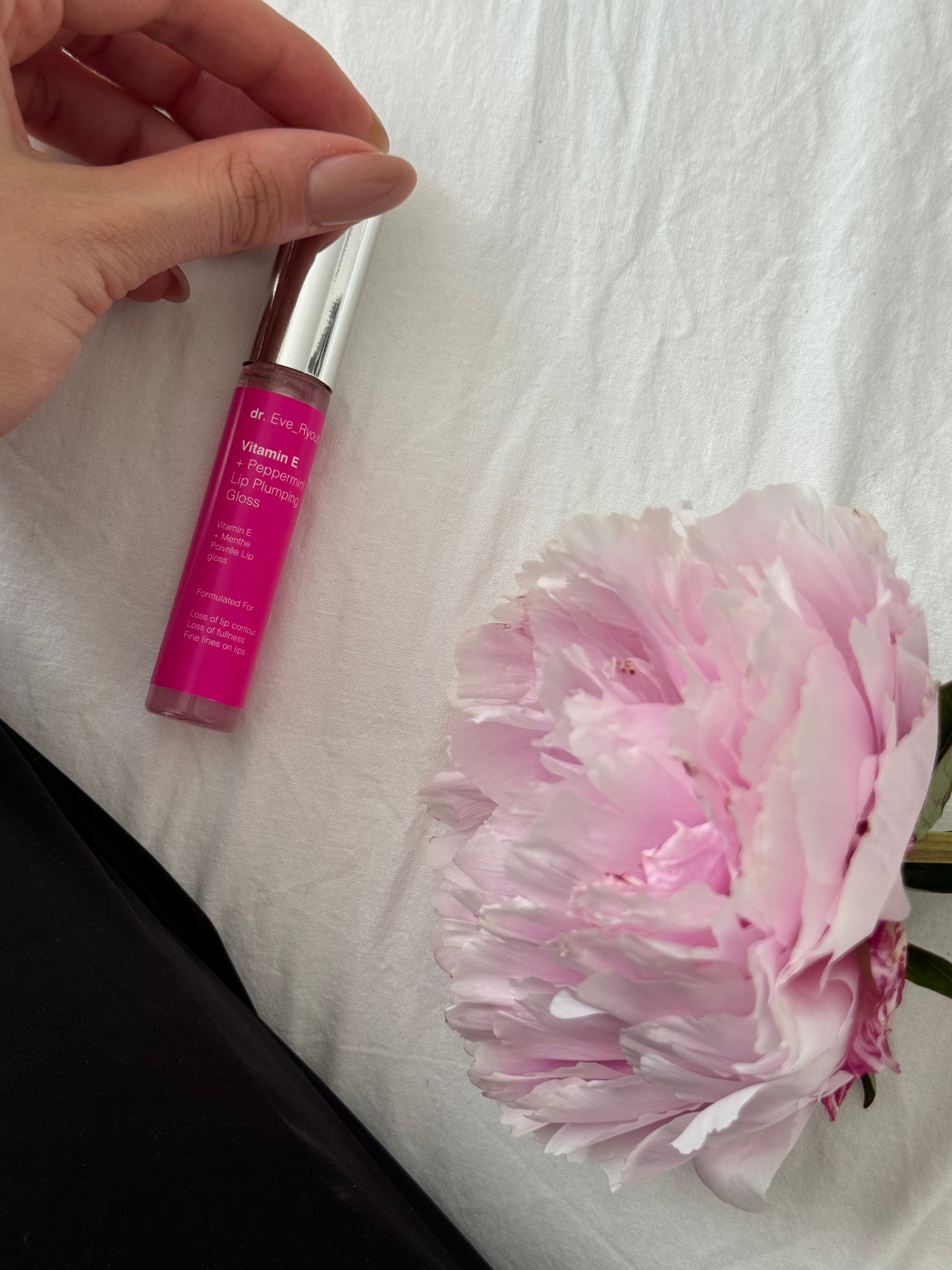 Un tube de gloss à côté d'une fleur. Liquide rose dans un contenant transparent avec un bouchon argenté. Inscription : Dr. Eve_Ryouth, Vitamine E + Menthe poivrée.