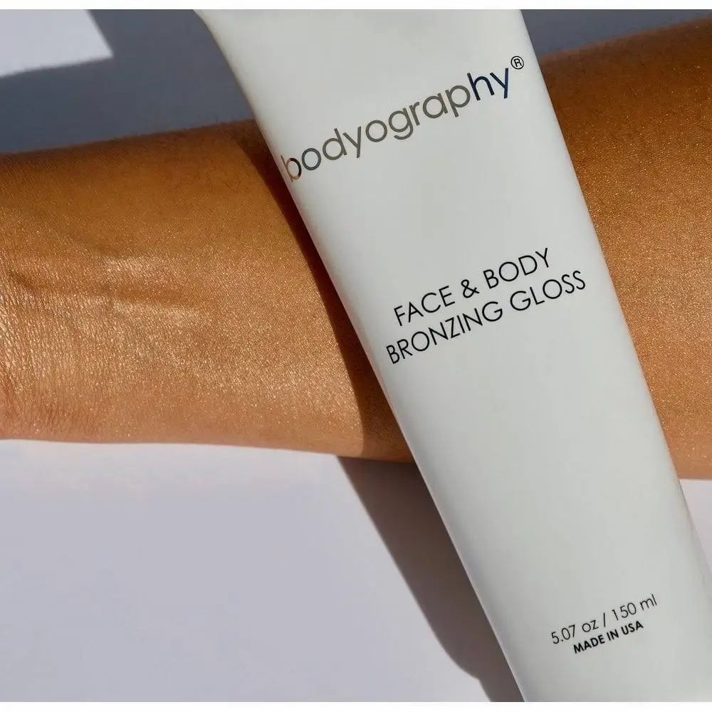 Witte tube met opschrift BODYOGRAPHY FACE & BODY BRONZING GLOSS. Aangebracht op arm. Inhoud: 5.07 oz / 150 ml.