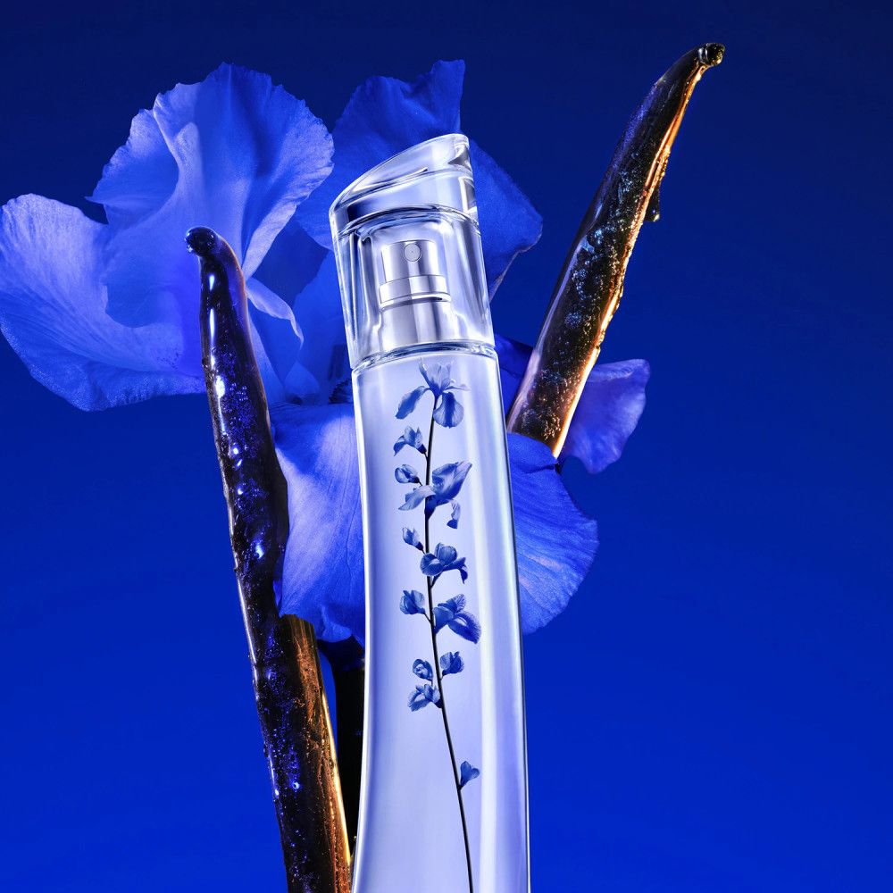 Flacon de parfum avec motif floral bleu, entouré de fleurs bleues et de bâtonnets bruns. Fond bleu.