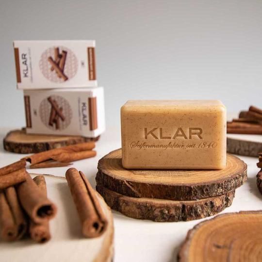 Zeep op een houten schijf, omringd door kaneelstokjes en verpakkingen. Zeep met opschrift KLAR. Verpakking met kaneelillustratie en tekst KLAR.