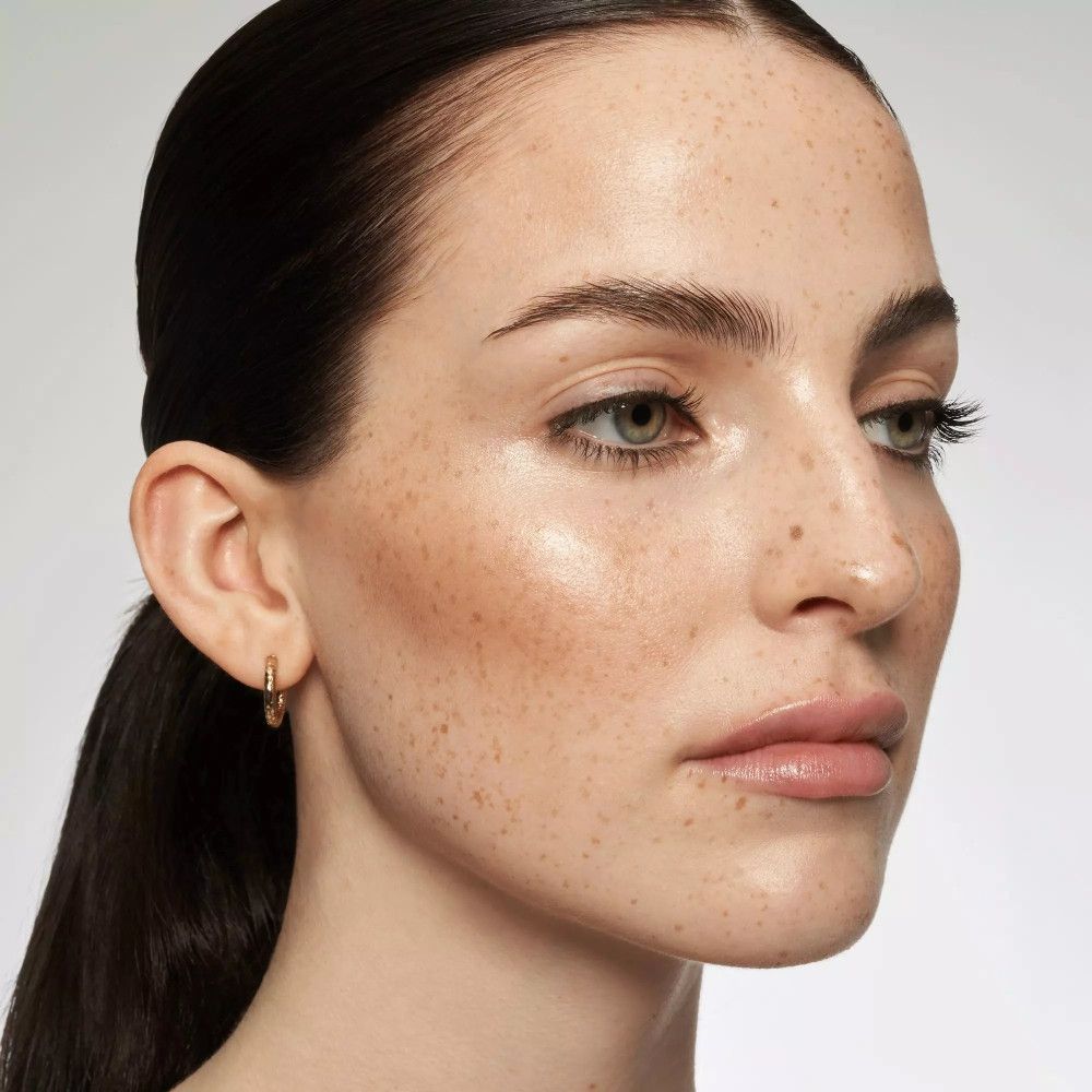 Gros plan d'un visage avec effet brillant. Peau avec des taches de rousseur. Créoles dorées. Cheveux foncés.