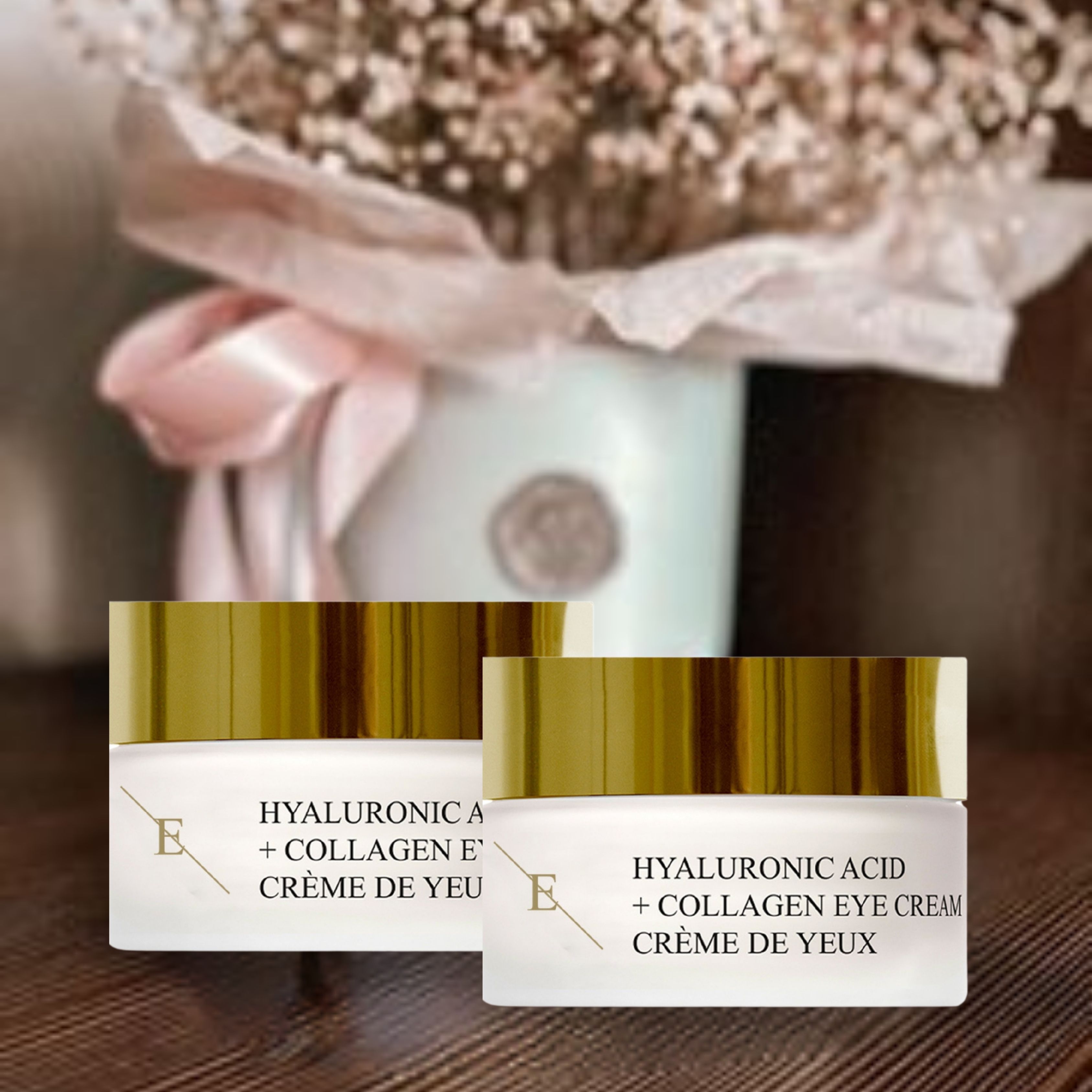 Twee potjes oogcrème met gouden deksels. Opschrift: Hyaluronzuur + Collageen Eye Cream. Crème de Yeux. Logo 'E'. Op een tafel, met bloemen op de achtergrond.