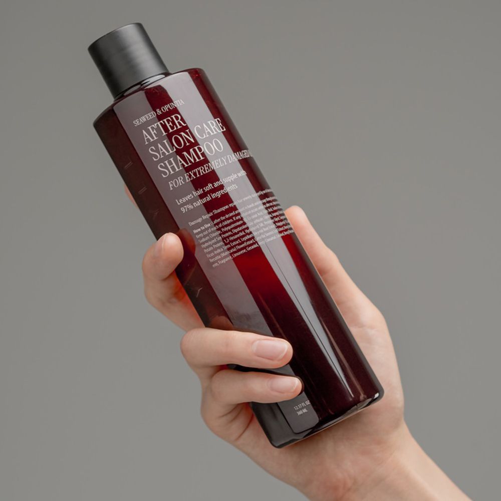 Hand houdt een bruine shampoo fles met zwarte dop. Opschrift: After Salon Care Shampoo voor extreem beschadigd haar. Label.