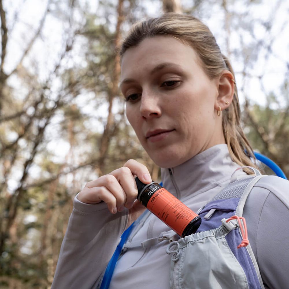 Femme avec sac à dos. Elle sort une bouteille OLEUS d'une poche. Forêt en arrière-plan.