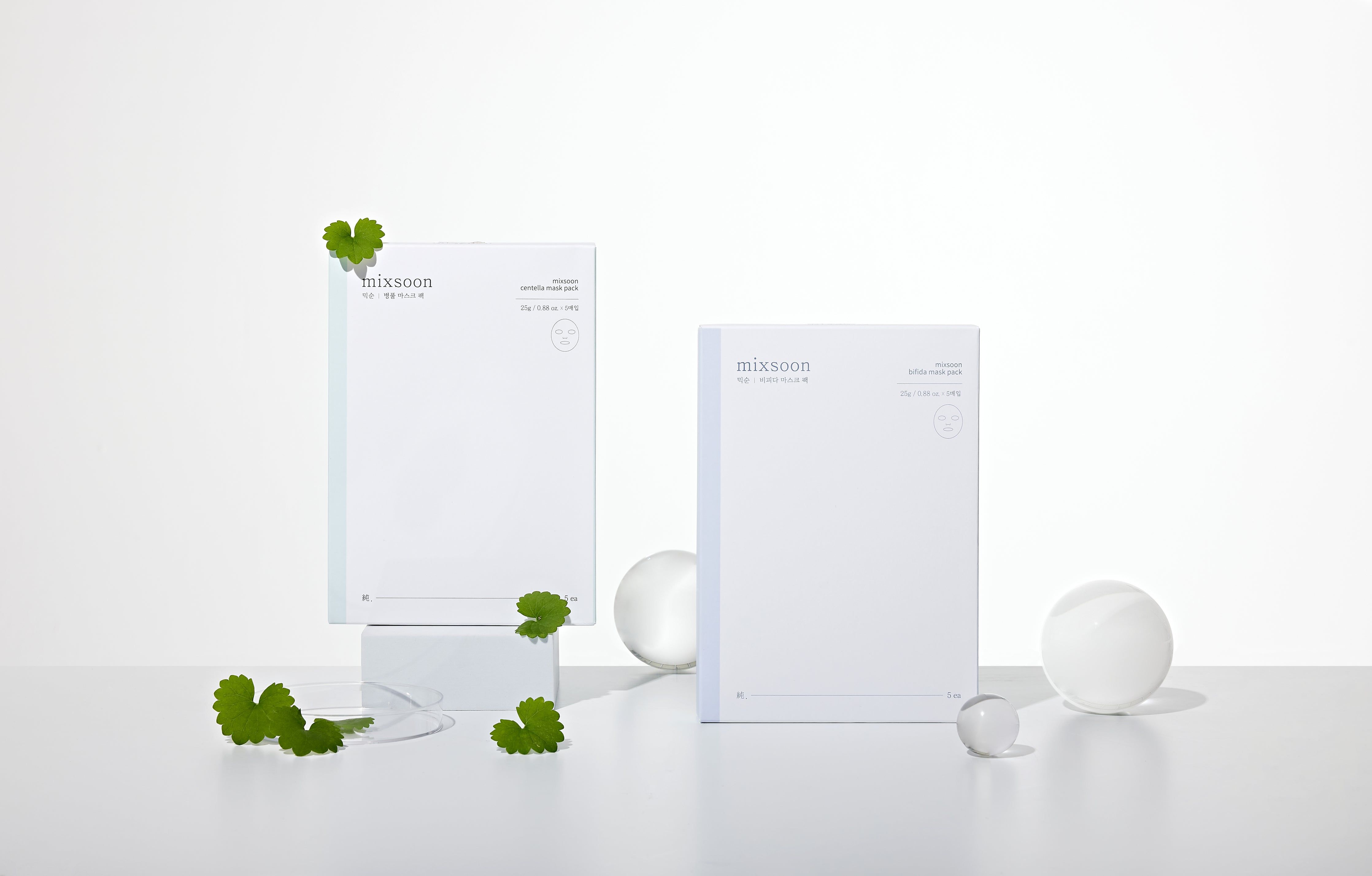 Deux emballages Mixsoon Centella Mask Pack. Paquets blancs rectangulaires. À côté, feuilles vertes et boules transparentes.