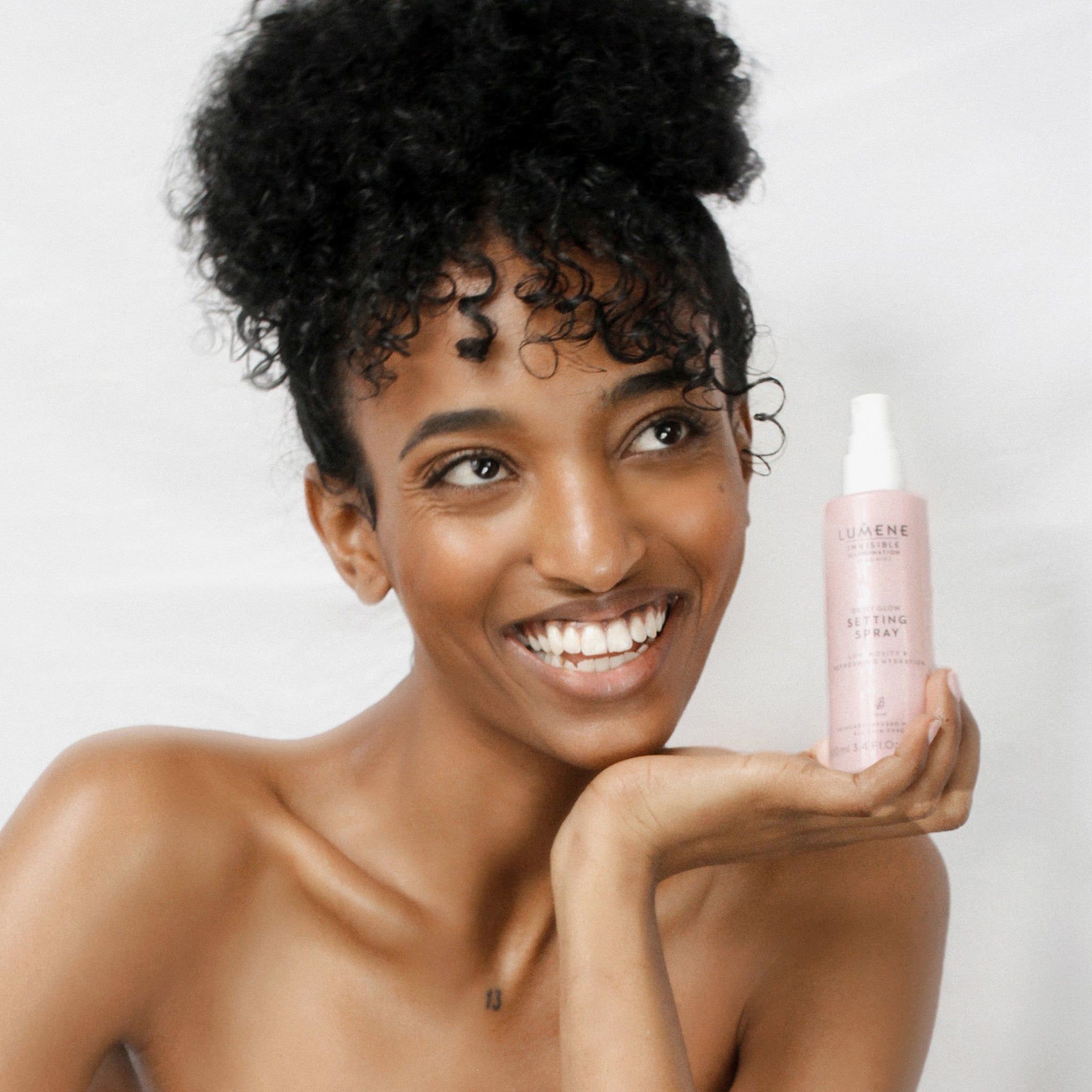 Vrouw houdt roze sprayfles vast. Opschrift: Lumene INVISIBLE ILLUMINATION Dewy Glow Setting Spray. Vrouw lacht. Achtergrond: wit.