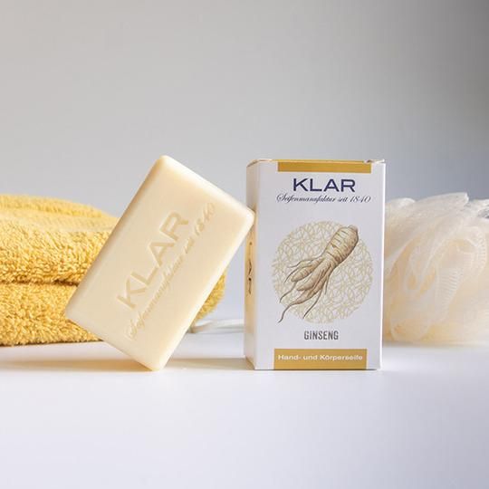 Zeep en verpakking naast handdoek en spons. Zeep met KLAR reliëf. Verpakking: KLAR, Ginseng, hand- en lichaamszeep.
