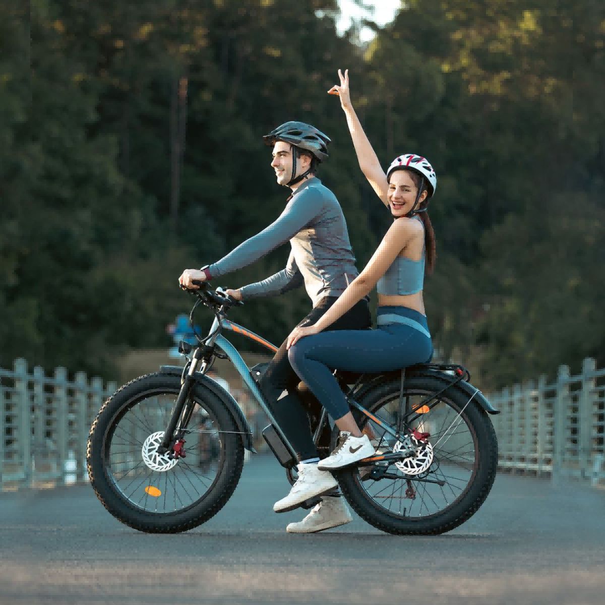 Twee personen rijden op een grijze elektrische fatbike. Ze dragen helmen. Achtergrond: bomen en brug.