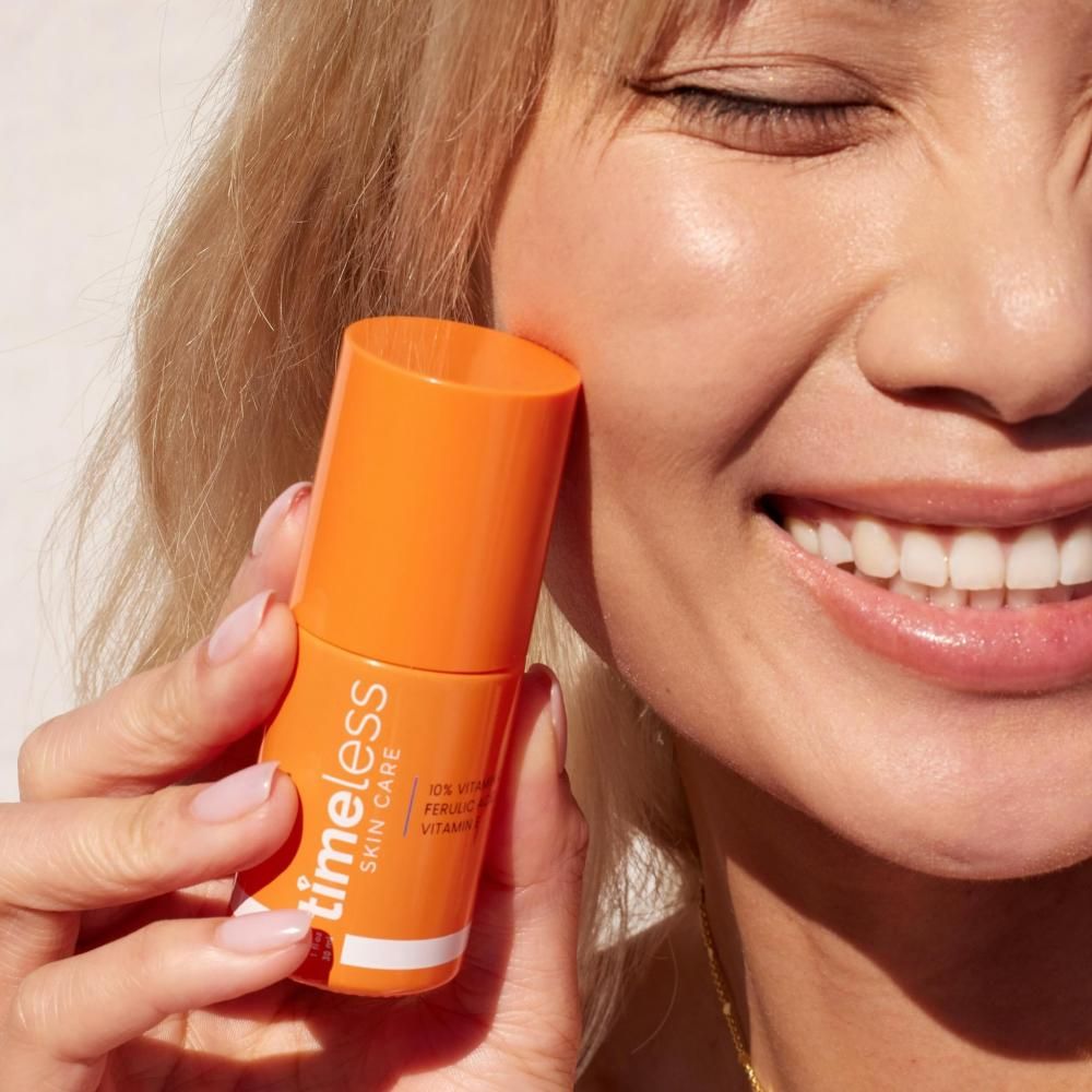 Femme tenant un flacon orange. Inscription : TIMELESS SKIN CARE, 10% Vitamine C, Acide férulique, Vitamine E. La femme sourit.
