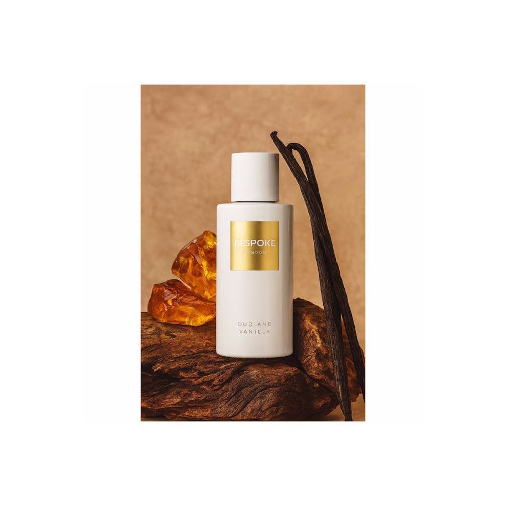 Flacon de parfum blanc avec étiquette dorée. Texte: Bespoke, Oud and Vanilla. Sur bois et gousses de vanille.