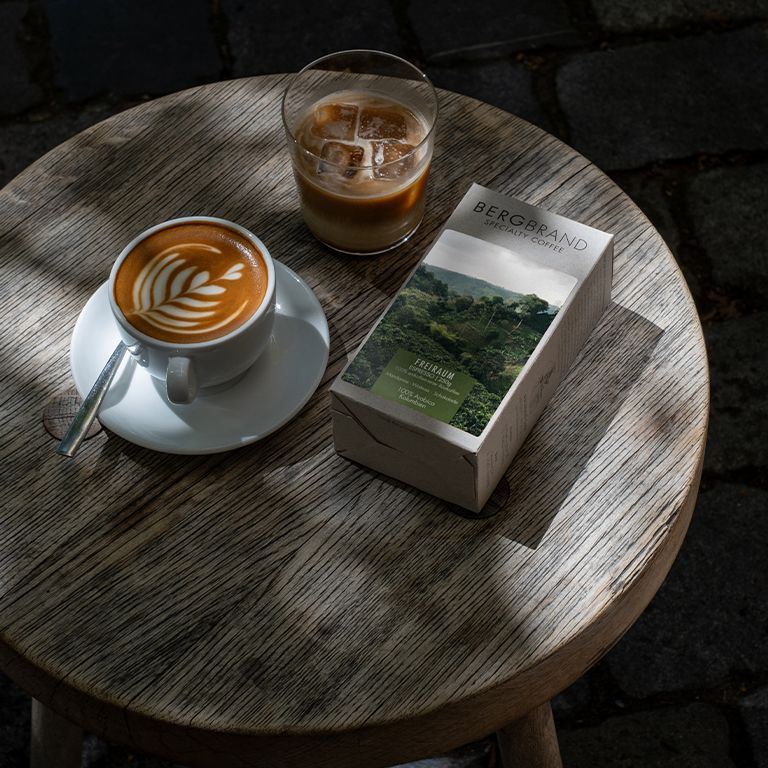 Café avec latte art et un verre de café glacé. À côté, un paquet de café. Sur une table en bois.