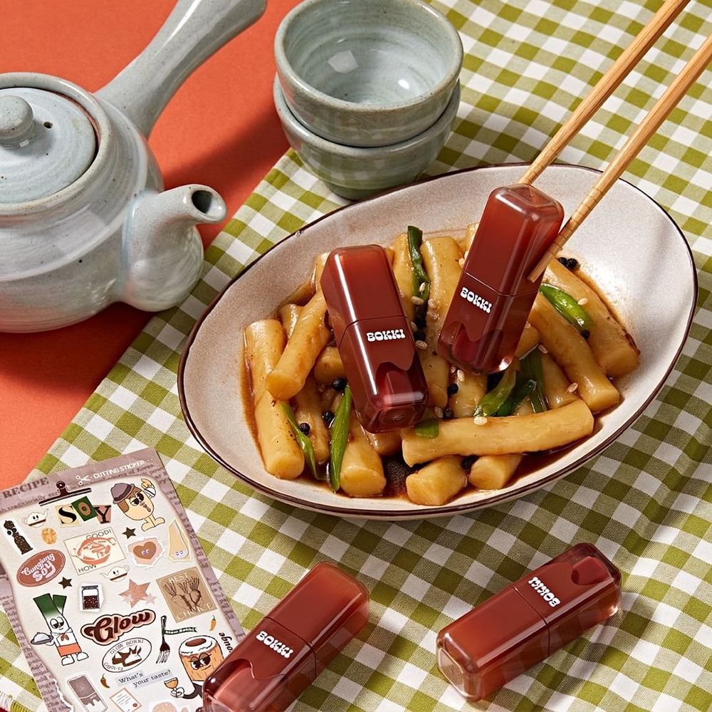 Trois gloss à lèvres sur un plat de nourriture. Deux sont tenus avec des baguettes. Inscription : BOKKI.