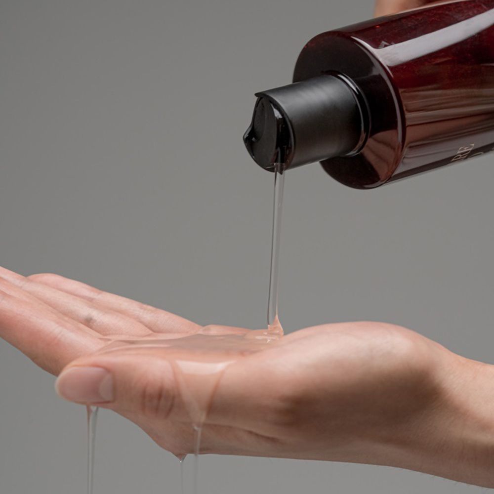 Hand giet shampoo uit een bruine fles. Vloeistof stroomt in de handpalm. Zwarte dop.