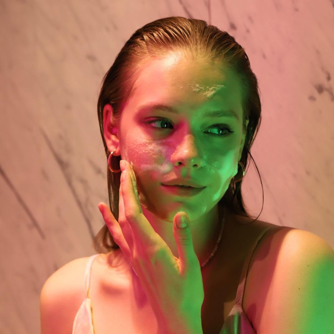 Visage d'une femme avec de la mousse sur la peau. Elle se touche le visage. Fond: mur éclairé en couleur.