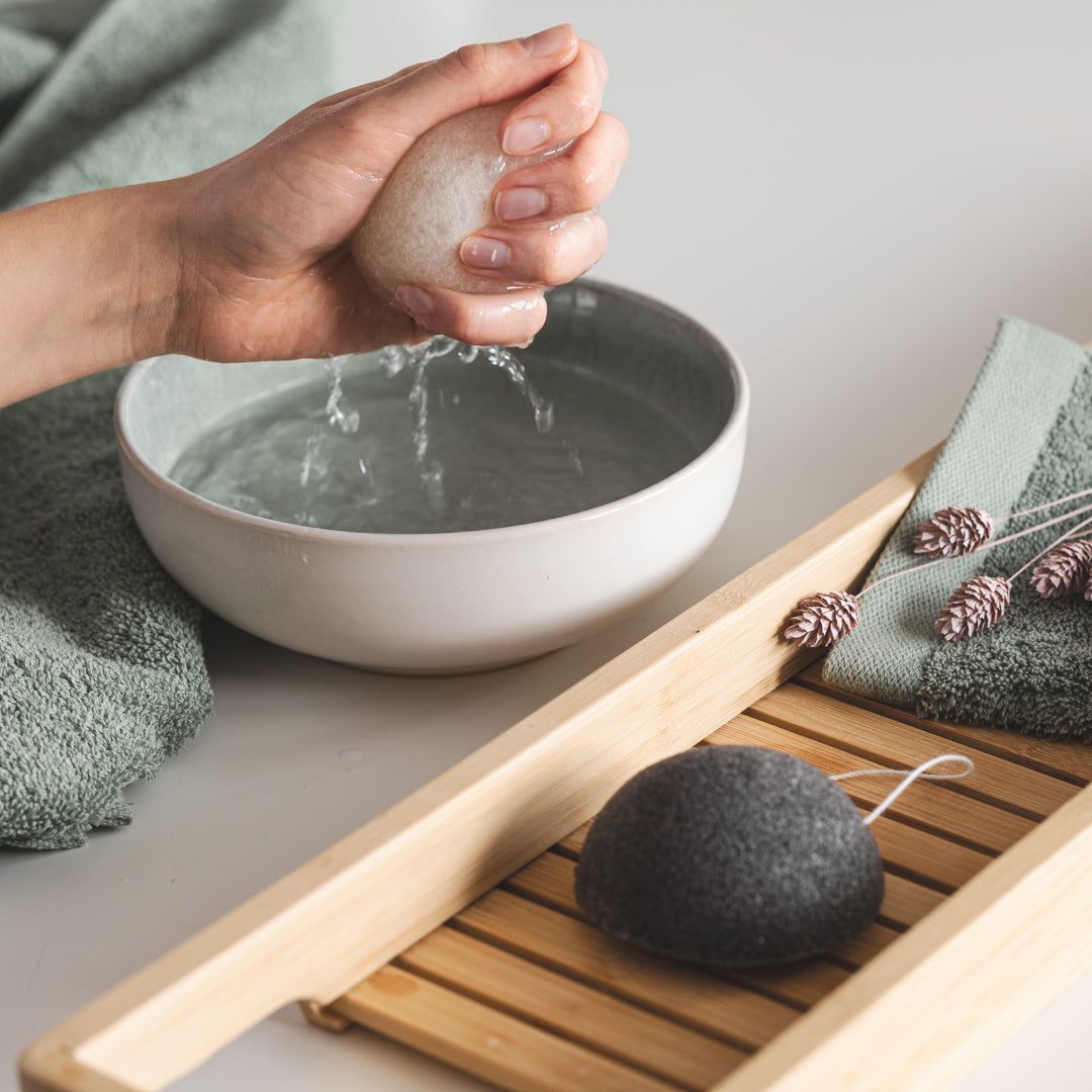 Une main presse de l'eau d'une éponge Konjac grise. En arrière-plan, un bol d'eau, un plateau et une serviette.