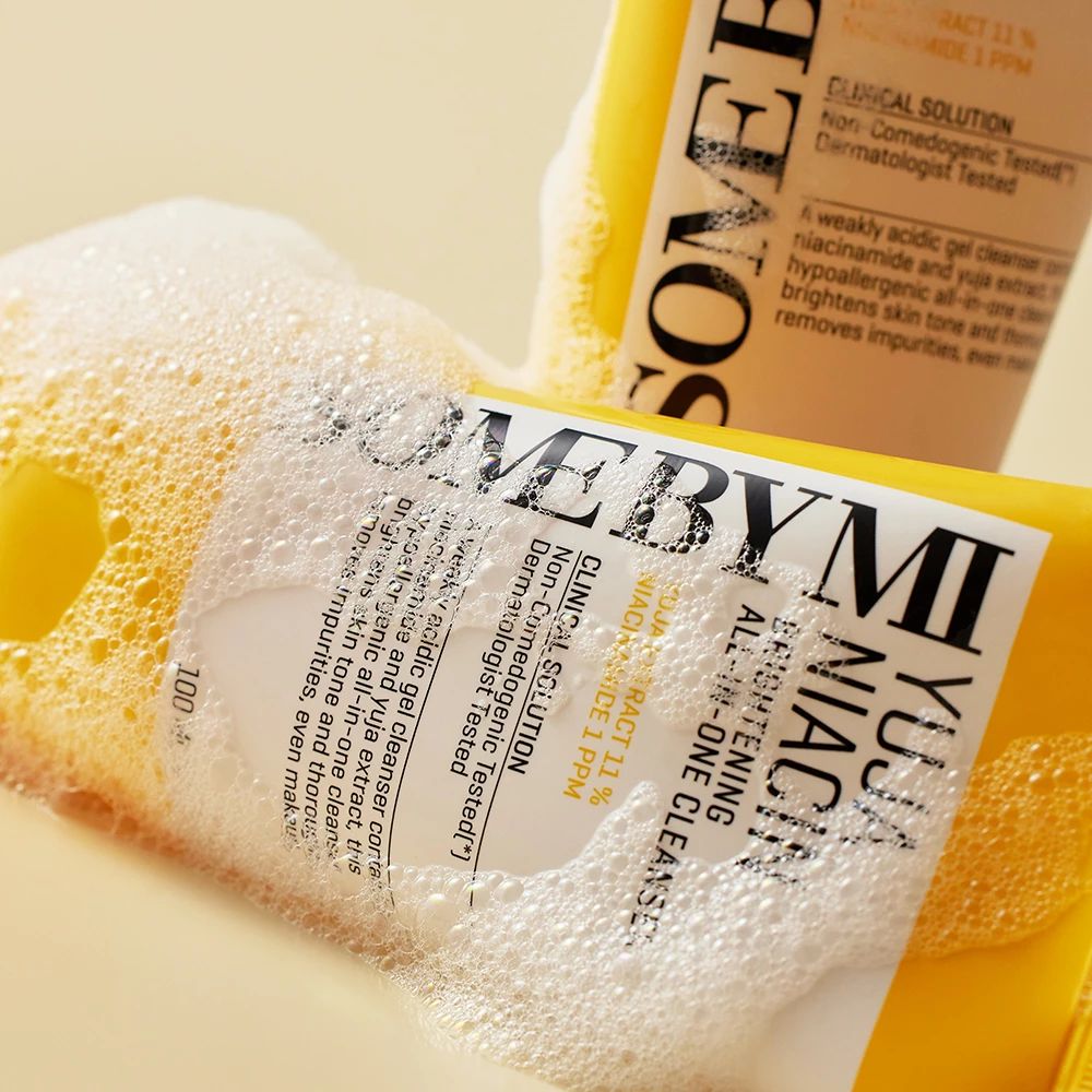 Gele tube met opschrift Yuja Niacin Brightening All-in-One Cleanser, omgeven door schuim. Tekst: Dermatologisch getest.